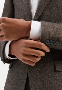 Brązowa marynarka o fakturze z czarnymi guzikami, białym mankietem wystającym spod rękawa. Zbliżenie na ręce poprawiające mankiet, ukazujące szczegóły tkaniny.