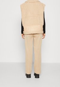 Person, der mit dem Rücken zur Kamera steht, trägt eine beige Sherpa-Weste, beige Hose, schwarze Langarmshirt und schwarze klobige Stiefel vor einem schlichten Hintergrund.