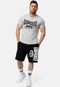 Pilkas TAPOUT marškinėliai su juodais užrašais, derinami su juodomis šortais, turinčiais baltą TAPOUT logotipą. Aprangą užbaigia baltos sportbačiai ir Dryžuotos kulkšninių kojinės.