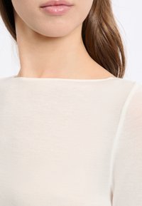 Top beige chiaro realizzato in un tessuto morbido ed elastico. Presenta un collo rotondo e maniche lunghe, con una texture morbida e trasparente.