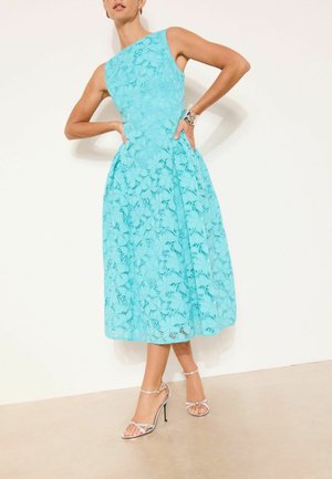 Žena v tyrkysovomodrých čipkovaných šatách bez rukávov, dĺžky po kolená, a strieborných sandáloch na vysokom podpätku stojí s rukami v bok proti bielej stene.