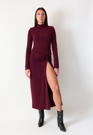 Abito bordeaux a maniche lunghe con uno spacco alto, caratterizzato da una scollatura a collo alto e un dettaglio arricciato sul lato, abbinato a stivaletti neri.