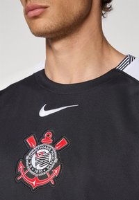 Tricou sport negru, cu un logo Nike alb. Tricoul are un material texturat și un emblemă detaliată în roșu și negru pe piept.