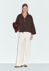Brun, løs bluse med V-hals og snøre detalje foran, parret med hvide brede bukser. Modellen holder en rød tekstureret clutch.