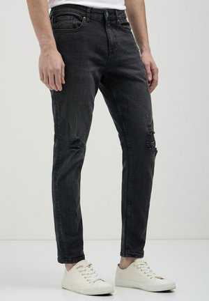 Jean skinny en denim noir avec des détails déchirés aux genoux, cinq poches et une taille régulière, associés à des baskets blanches.