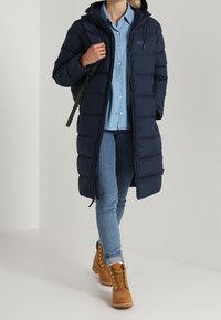 Marinblå pufferjacka med huva, dragkedjestängning, sidofickor och kviltad design. Bärs över en ljusblå skjorta och jeans, i kombination med beige stövlar.