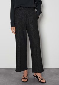 Zwarte wijde broek met een gestructureerde, sprankelende afwerking. Heeft een hoge taille en plooien aan de voorkant. Gemaakt in combinatie met zwarte sandaaltjes met banden.