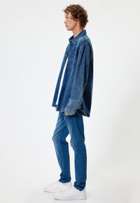 Camisa de mezclilla en azul oscuro con bolsillos delanteros, tejido superpuesto y cuello; combinada con jeans azul claros y zapatillas blancas.