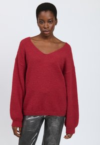 Maglione rosso voluminoso con scollo a V e maniche lunghe e larghe, realizzato in un morbido materiale texturizzato; indossato con pantaloni metallici grigi lucidi.