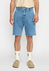 Lichtblauwe denim shorts met een ontspannen pasvorm, hebben een standaard tailleband, opgerolde zomen en zichtbare stiksels. De shorts worden gedragen met crèmekleurige sokken en pantoffels.