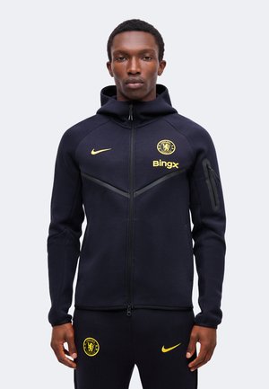 Uomo che indossa felpa con cappuccio e zip blu scuro Nike Chelsea FC e pantaloni abbinati con loghi gialli, in piedi davanti a uno sfondo chiaro uniforme.