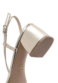 Sandale à talons hauts en or métallique avec un bout carré, une fine bride à la cheville et une semelle en caoutchouc texturé pour une meilleure adhérence. Détail avec un logo sur la semelle.