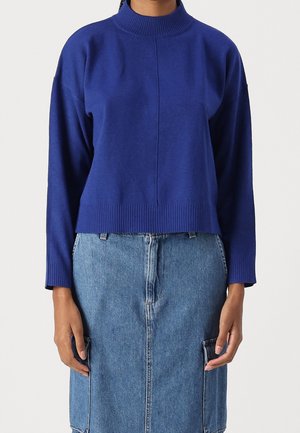Femme portant un pull à manches longues bleu roi avec des poignets et un ourlet côtelés, associé à une jupe en denim bleu mi-longue avec de grandes poches avant.