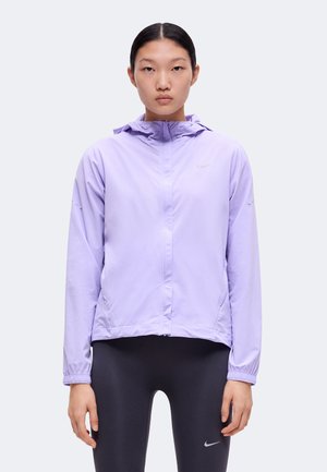Jeune femme aux cheveux noirs attachés, portant une veste zippée lilas et des leggings noirs avec un petit logo Nike sur chacun