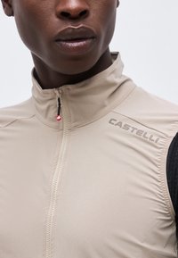 Nahaufnahme einer Person, die eine beige ärmellose Zip-up-Weste mit dem "Castelli"-Logo auf der rechten Schulter trägt.