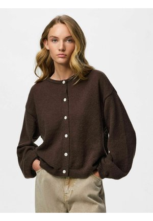 Jeune femme aux cheveux blonds ondulés portant un cardigan marron foncé boutonné et un pantalon beige clair, les mains dans les poches.