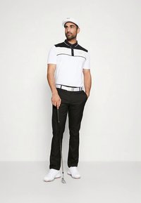 Άνδρας με άσπρη και μαύρη γκολφ στολή κρατώντας ένα γκολφ κλαμπ, στέκεται με το ένα χέρι στην τσέπη σε έναν απλό φόντο.