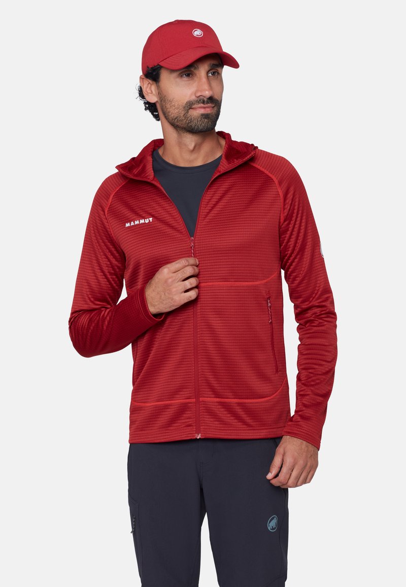 Uomo con giacca rossa con cerniera e cappuccio e cappellino rosso coordinato, che aggiusta la cerniera, in piedi davanti a uno sfondo chiaro.