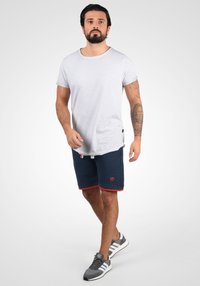 Grijze katoenen t-shirt met een ronde hals, gecombineerd met marineblauwe shorts met rode accenten en een logo. Grijze sneakers maken de outfit af.
