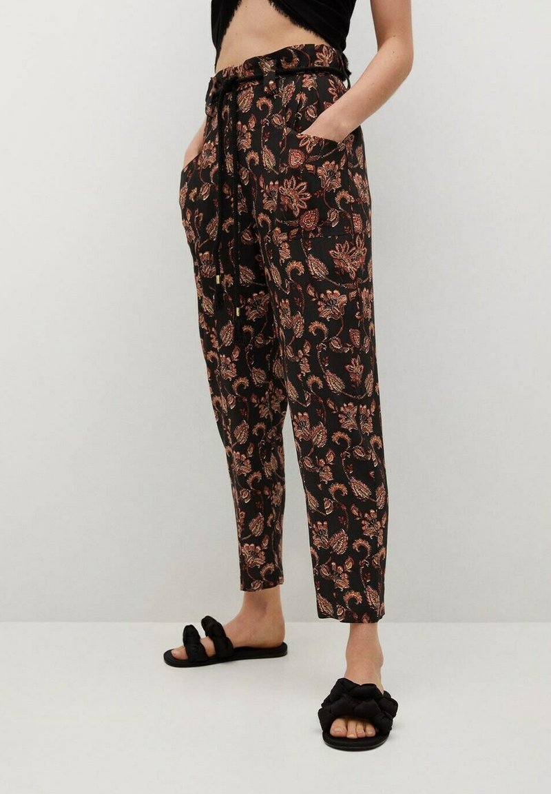 Personne portant un pantalon noir à motif floral avec poches, taille nouée, et des sandales noires à brides tressées, debout devant un fond uni.