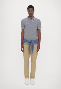 Camisa polo a rayas para hombre en azul marino y blanco con collar, combinada con pantalones cargo beige y zapatillas blancas. Suéter azul atado a la cintura.