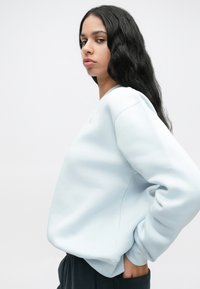 Νεαρή γυναίκα με μακριά μαύρα μαλλιά, φορώντας ένα ανοιχτό μπλε φούτερ και μαύρο παντελόνι, στέκ condensed 1 με το ένα χέρι στην τσέπη της, μπροστά από έναν απλό φόντο.