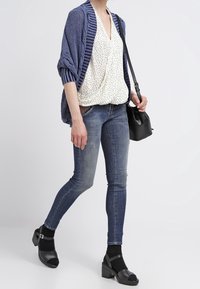 Cardigan de punto azul, blusa blanca de lunares y jeans oscuros ajustados. Sandalias negras con tacón cuadrado y un pequeño bolso cruzado negro.