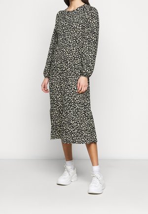 Kobieta w czarno-białej midi sukience w cętki lamparta z długimi rękawami i białych trampkach stojąca na jednolitym tle.