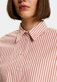 Camicia a righe rosse e bianche con colletto a punta, due tasche anteriori e chiusura con bottoni. Il tessuto sembra morbido e leggero.