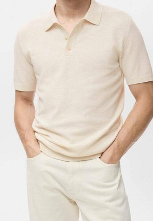 Homme portant un polo tricoté beige à manches courtes avec des boutons, rentré dans un pantalon beige taille haute, mains dans les poches, fond neutre.