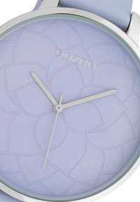 Montre ronde avec un boîtier en métal argenté et un bracelet en cuir bleu clair. Le cadran lavande présente un motif floral et des aiguilles argentées, avec le texte "OOZOO".