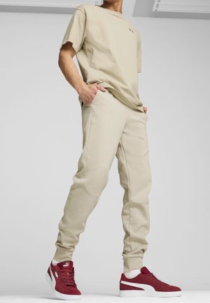 Person wearing beige casual pants and matching t-shirt with red and white Puma sneakers, standing with hands in pockets.