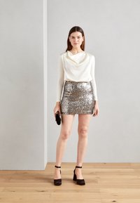 Weißes Oberteil mit Schalkragen und langen Ärmeln, kombiniert mit einem silbernen Pailletten-Minirock. Schwarze Wedge-Heels mit Knöchelriemen und eine kleine schwarze Clutch. Holzboden als Hintergrund.