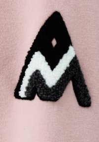 Rosa tyg med en texturerad broderad logotyp, med svarta, vita och grå färger i en geometrisk design som liknar en abstrakt bokstav.