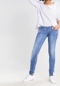 Jeans ajustados azul claro con un lavado desgastado. Combinados con una camiseta de manga larga a rayas y holgada. Completado con zapatillas blancas.