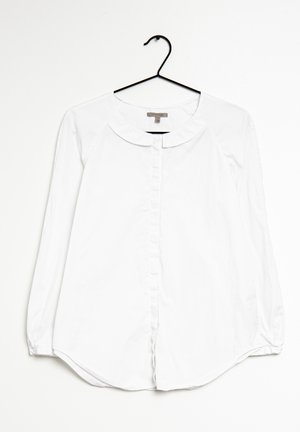 Blouse blanche à manches longues avec boutons et col arrondi suspendue à un cintre noir sur fond blanc.