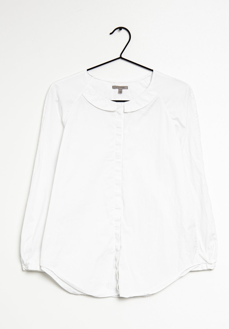 Blouse blanche à manches longues avec boutons et col arrondi suspendue à un cintre noir sur fond blanc.
