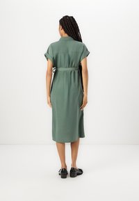 Vestito midi verde con colletto, maniche arrotolate e dettaglio in vita legato. Tessuto liscio e taglio dritto, abbinato a scarpe nere.