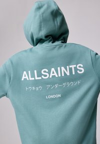 Türkiz kapucnis pulóver húzózsinórral és egy nagy fehér grafikával a hátán, amelyen az "ALLSAINTS" felirat és japán szöveg, valamint a "LONDON" szerepel.