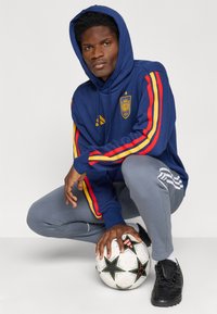 Ναυτικό φούτερ με κόκκινες και κίτρινες ρίγες, έμβλημα Ισπανίας, γκρι φόρμες, μαύρα υφασμάτινα ποδοσφαιρικά παπούτσια, κρατώντας μία μπάλα ποδοσφαίρου με αστέρια σε μαύρο και άσπρο.