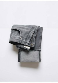 Jean en denim gris plié avec une ceinture visible, un bouton, une passoire de ceinture et une étiquette de vêtement blanche sur une surface blanche.