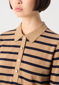 Primo piano di una persona che indossa una polo a righe beige e blu navy con quattro bottoni dorati e capelli scuri corti.