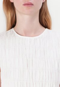White, textured top with horizontal pleats and a round neckline, displaying soft, lightweight fabric. Features a smooth finish and minimalist design.