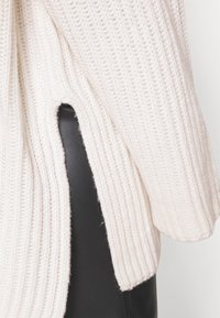 Crème-kleurige gebreide trui met geribbelde textuur, voorzien van een zijsplit, gecombineerd met zwarte stof eronder.
