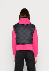 Schwarze gesteppte Weste über einem rosa Kapuzenpullover. Die Weste zeichnet sich durch ein kurzes Design mit einer glatten Textur und gestepptem Rautenmuster aus.