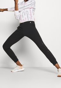 Pantaloni da jogging neri atletici con un design aderente, dotati di polsini elasticizzati e un delicato dettaglio del logo lungo il lato. Abbinati a sneaker chiare.