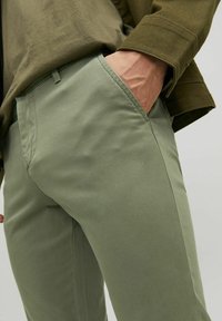 Pantalons en coton vert sur mesure avec une texture lisse, poches standards et coupe décontractée, associés à une chemise marron à la taille.