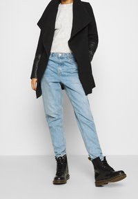 Abrigo de lana negro con mangas de cuero, suéter de punto gris, jeans de mezclilla azul claro y botas negras con cordones y suela gruesa.