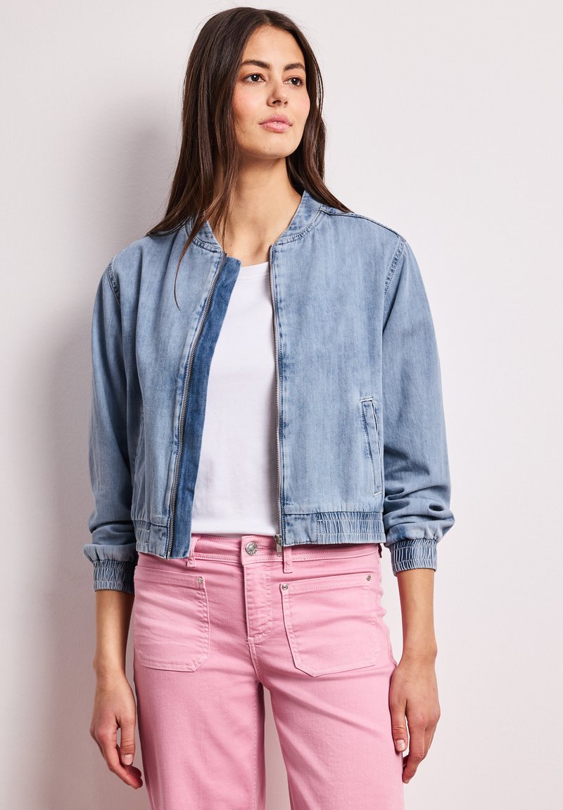 Woman wearing light blue denim jacket over white shirt and pink pants, standing against plain light background.