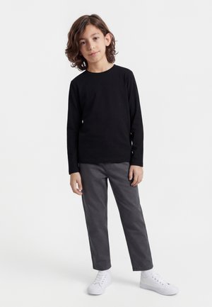 Chłopiec z brązowymi włosami do ramion, ubrany w czarną koszulę z długim rękawem, szare spodnie i białe tenisówki, stojący na białym tle.